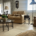 A well-lit living room with beige sofas, a glass coffee table, a side table with a lamp, and a wall clock. Plants decorate the coffee table and a side table by the window with shutters.
